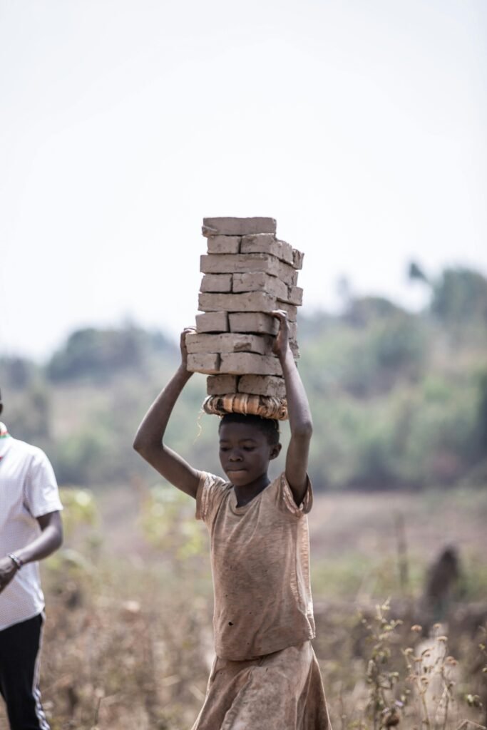 A young boy carries a stack of bricks outdoors in a rural setting, documenting daily life.